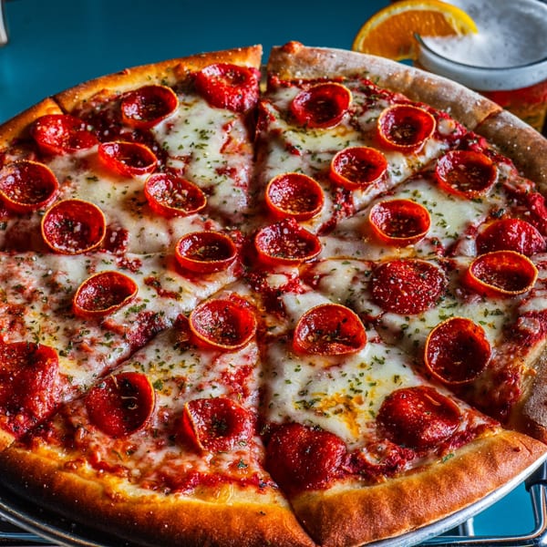 A pepperoni pizza with melted cheese and herbs, with a beer and orange slice in the background.
