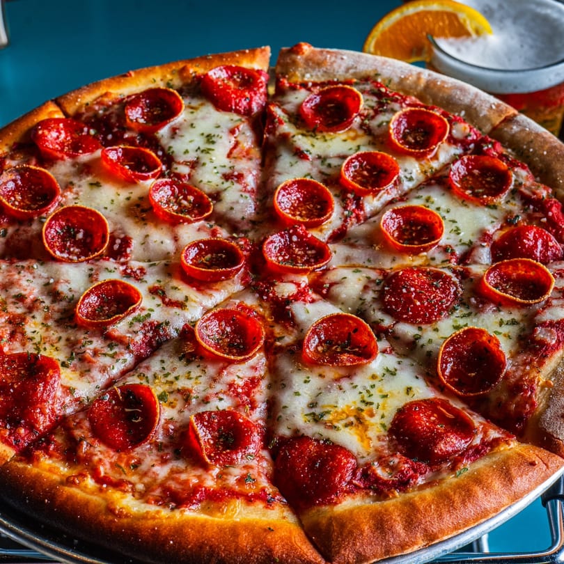 A pepperoni pizza with melted cheese and herbs, with a beer and orange slice in the background.